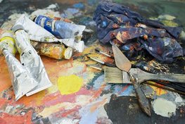 Auf einer farbverschmierten Palette liegen mehrere Farbtuben, Pinsel, ein kleiner Spachtel sowie ein Lappen mit Farbklecksen.